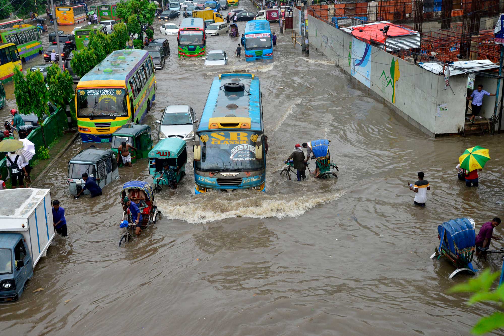 Buses, cars and bicycles attempt to travel on a heavily flooded road, which reaches people's waists.