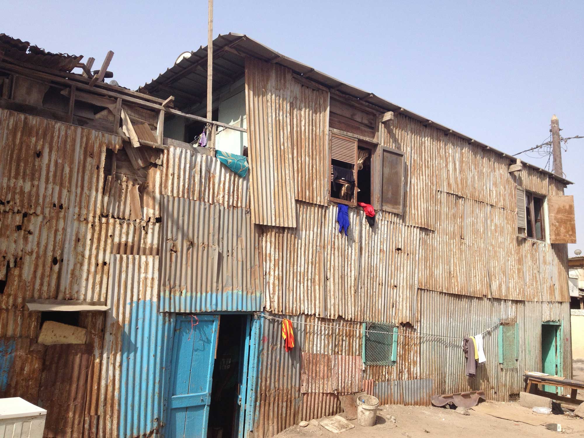 A shack made of corrugated metal that is beginning to rust in the intense heat. Lines of clothes that are drying in the sun are visible around the house and the doors are painted bright green and blue.