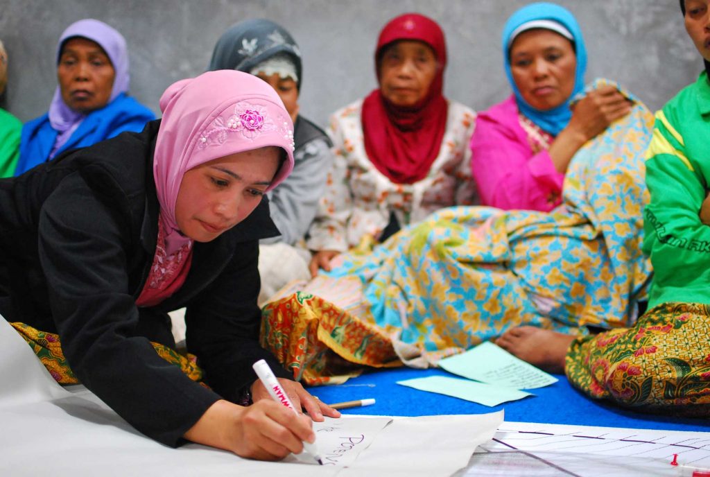 A group of women participating in a planning session, with one lent over and writing down notes on paper while the others overlook.